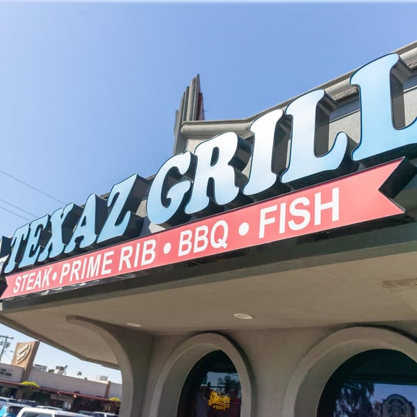 Texaz Grill sign with "Steak, Prime Rib, BBQ, Fish" on a red banner, under a blue sky.