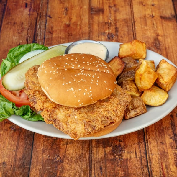 Crispy fried chicken sandwich with lettuce, tomato, pickle, and roasted potatoes on a white plate.