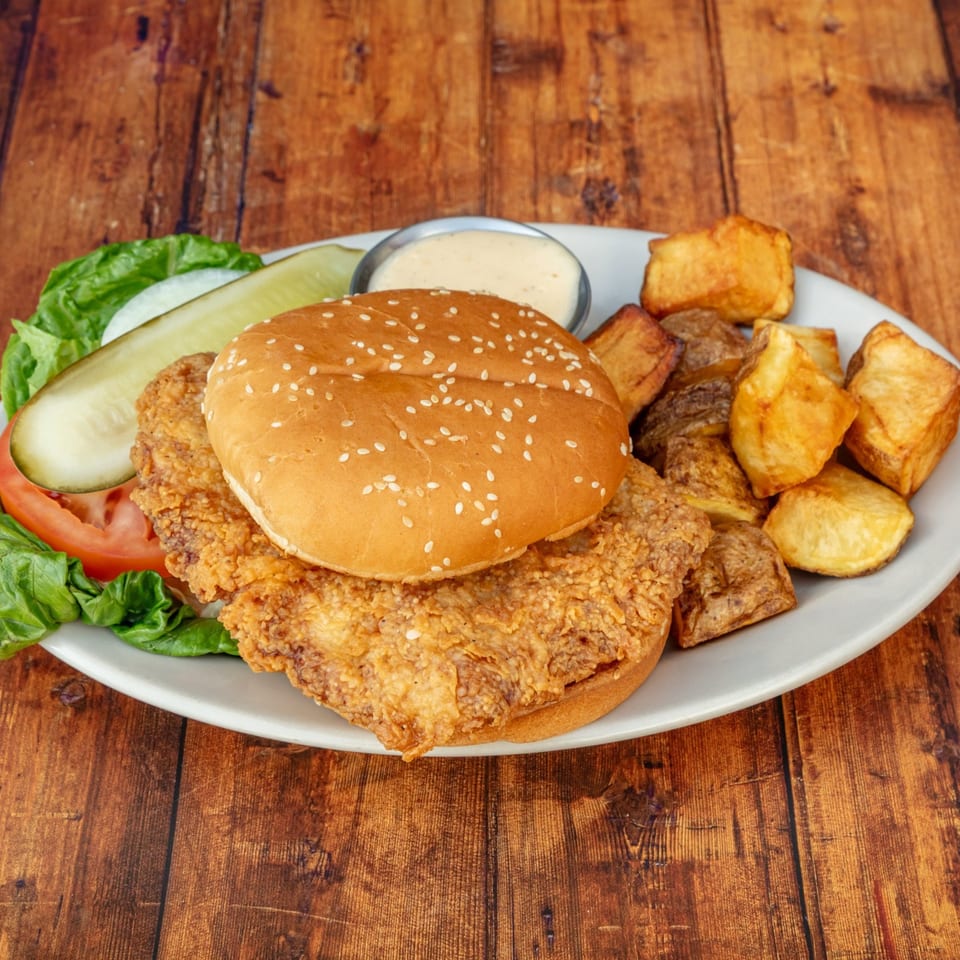 Crispy fried chicken sandwich with lettuce, tomato, pickle, and roasted potatoes on a white plate.