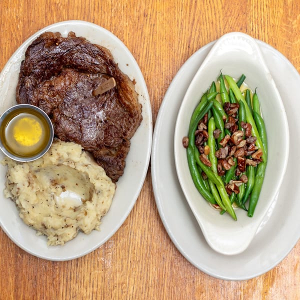 A medium-cooked steak with mashed potatoes, melted butter, and a side of green beans with pecans.