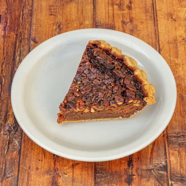 A slice of pecan pie with a flaky crust sits on a white plate on a wooden table.