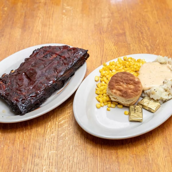 Plates of BBQ ribs, corn, mashed potatoes with gravy, a biscuit, and butter on a wooden table.
