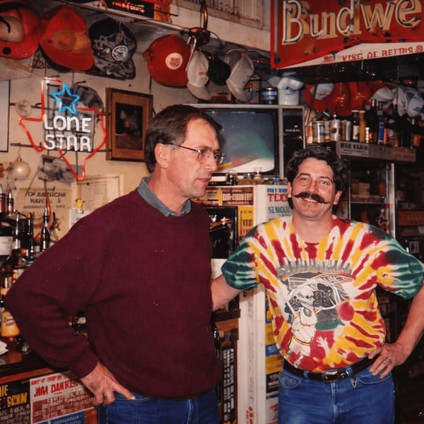 Two men standing in a rustic bar with a Lone Star neon sign and Budweiser banner.